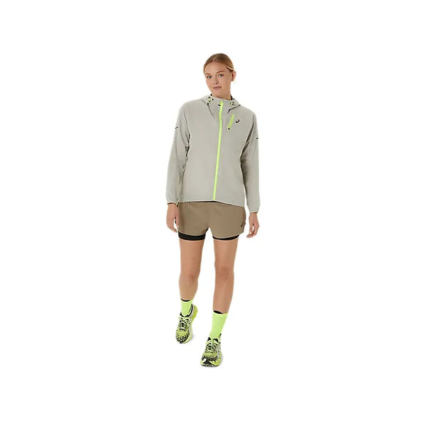 En person står på en hvid baggrund iført en lysegrå jakke med hætte og neongule detaljer, Asics Fujitrail 2-N-1 Shorts i Dame Pepper/Performance Black, neongule sokker og Performance Black/neongule sneakers.