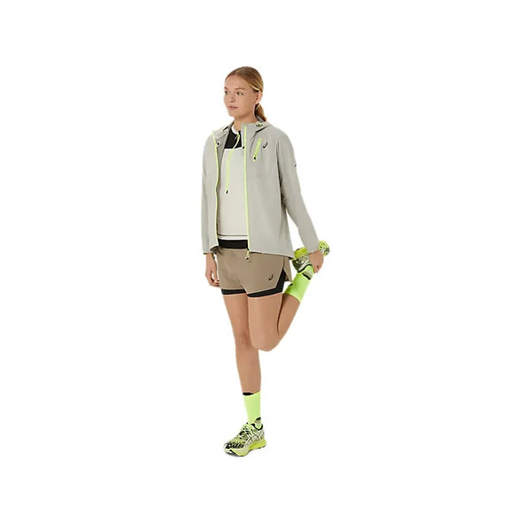 En person iført lysegrå jakke, Asics Fujitrail 2-N-1 Shorts til kvinder i Pepper/Performance Black, neongule sokker og neongule sneakers står på det ene ben og strækker det andet ved at holde anklen bag sig.