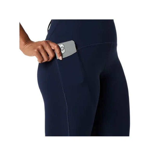 En person, der ses fra taljen og ned mod en hvid baggrund, lægger en smartphone i sidelommen på Asics Road High Waist Tight, Dame, Midnatsmørkeblå tights lavet af genbrugsmaterialer.