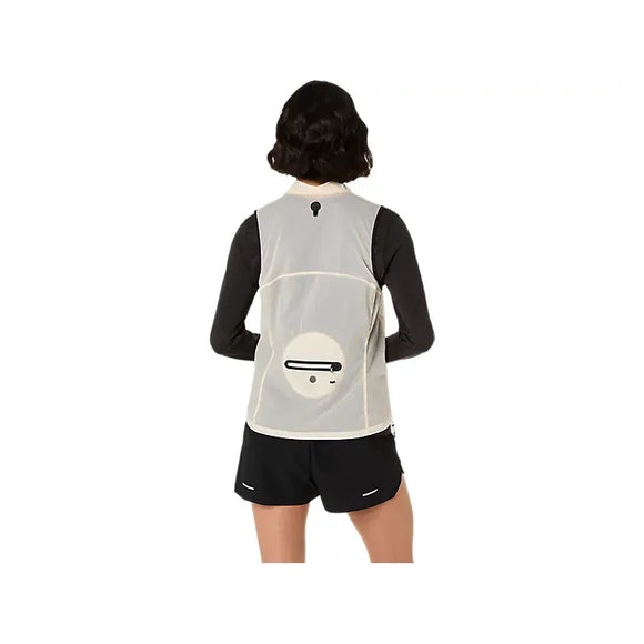 En kvinde med kort, mørkt hår ses bagfra iført en Asics Metarun Packable Gilet Dame i creme - en slank, vind- og vandafvisende løbevest med cirkulær lomme og sort ikon øverst på ryggen - over en langærmet sort skjorte og shorts.