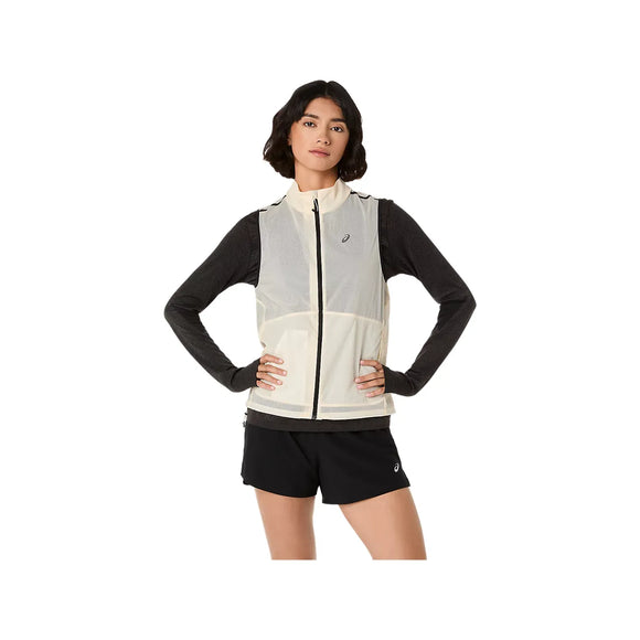 En kvinde står med ansigtet fremad og hænderne på hofterne, iført en Asics Metarun Packable Gilet, Dame, Cream over en langærmet mørk skjorte og sorte sportsshorts. Baggrunden er helt hvid.