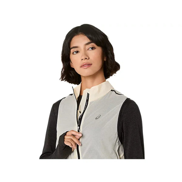 En kvinde med kort mørkt hår bærer Asics Metarun Packable Gilet, Dame, Cream - en vind- og vandafvisende vest - over en sort langærmet top. Hun kigger til siden, mens hun holder lynlåsen mod en almindelig hvid baggrund.