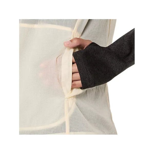 Et nærbillede viser en hånd i et mørkt langt ærme med hul til tommelfingeren, der glider ned i sidelommen på den cremefarvede Metarun Packable Gilet til kvinder fra Asics, der har et gennemsigtigt gittermønster og vind- og vandafvisende stof.