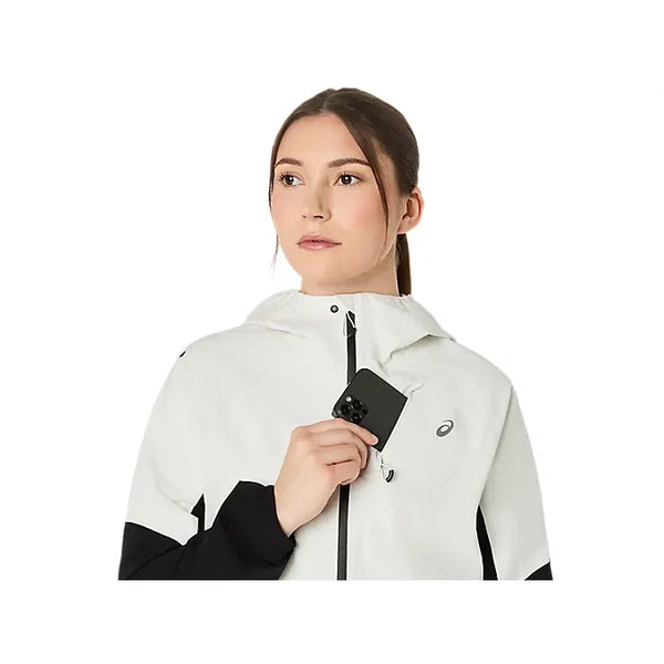 En kvinde i Asics Metarun Legend Jacket, Dame, Performance Black/Cream holder en sort smartphone ved brystet og kigger til siden mod en hvid baggrund - ideelt til dem, der vil have en stilfuld Asics løbejakke eller premium løbetøj.