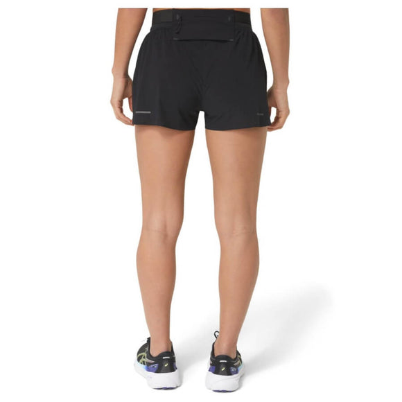 En kvinde står med ryggen til kameraet iført Asics Metarun Split Shorts, Dame, Performance Black og hvide løbesko med sorte og grønne detaljer. Baggrunden er helt hvid.