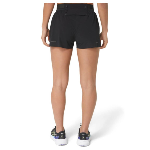 En kvinde står med ryggen til kameraet iført Asics Metarun Split Shorts, Dame, Performance Black og hvide løbesko med sorte og grønne detaljer. Baggrunden er helt hvid.