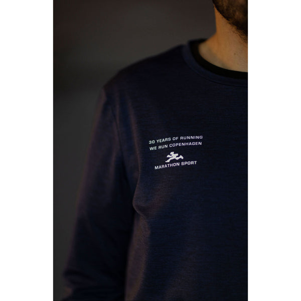 En person bærer Saysky Marathon Sport 30 Års Jubilæum Clean Pace Long Sleeve Unisex i blå med "WE RUN COPENHAGEN" i hvid tekst. Overkroppen og en del af ansigtet er synlige mod en mørk baggrund.