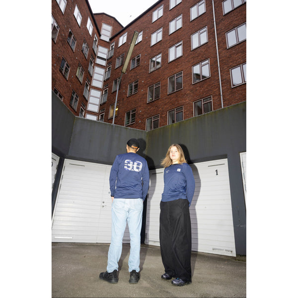 To personer står foran hvide garageporte med en murstenslejlighed bagved. Personen til venstre, klar til løbetur i København, er iført den blå Saysky Marathon Sport 30 Års Jubilæum Clean Pace Long Sleeve Unisex og lyse jeans; den anden ser fremad.