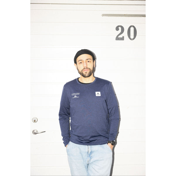 En skægget mand i sort hue, lyse jeans og blå Saysky Marathon Sport 30 Års Jubilæum Clean Pace Long Sleeve Unisex står foran en hvid dør mærket 20 med hænderne i lommerne og kigger ind i kameraet.