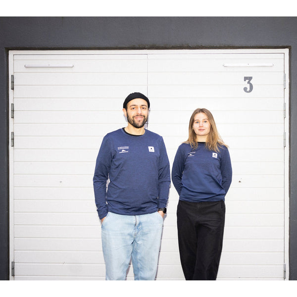 To personer står ved en hvid garageport mærket "3", begge iført blå Saysky Marathon Sport 30 Års Jubilæum Clean Pace Long Sleeve Unisex-trøjer. Den venstre person kombinerer den med lyse jeans og en sort hue, klar til løbetur København.