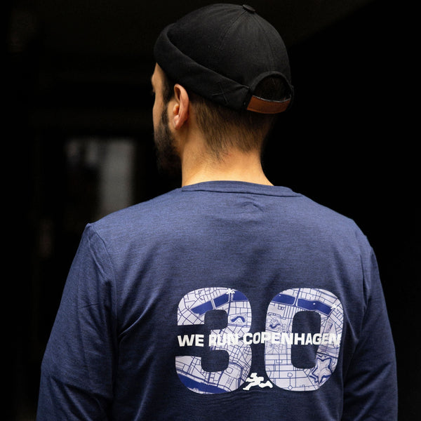 En person iført en blå Saysky Marathon Sport 30 Års Jubilæum Clean Pace Long Sleeve Unisex står med ryggen til kameraet og viser nummer 39 og "WE RUN COPENHAGEN", klar til løbetur i København.