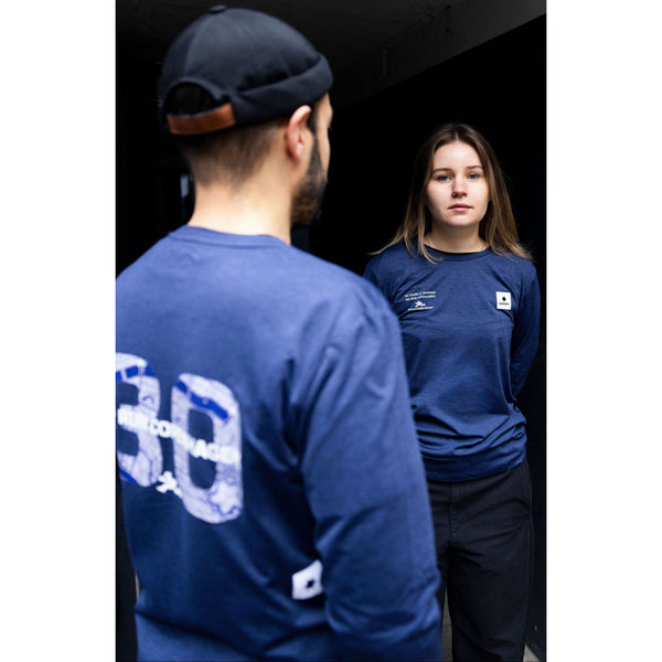 En mand og en kvinde er iført blå Saysky Marathon Sport 30 Års Jubilæum Clean Pace Long Sleeve Unisex-trøjer indendørs og står over for hinanden; manden ses bagfra, mens kvinden står over for kameraet med hænderne i lommerne.