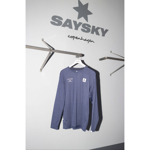 En blå Saysky Marathon Sport 30 Års Jubilæum Clean Pace Long Sleeve Unisex hænger på en hvid bøjle mod en lysegrå væg med SAYSKY-logoet og WE RUN COPENHAGEN over. Der er to tomme vægmonterede stativer på hver side.