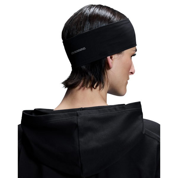 En person med kort, mørkt hår ses bagfra iført et sort On Core Headband, Unisex - mærket med ON på siden - og en sort jakke med hætte. Baggrunden er hvid.