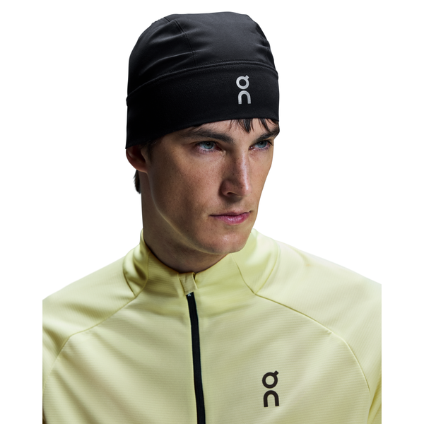 En person iført On Core Beanie i sort og en lysegul sportsjakke med lynlås og sort logo kigger ud til siden mod en hvid baggrund.