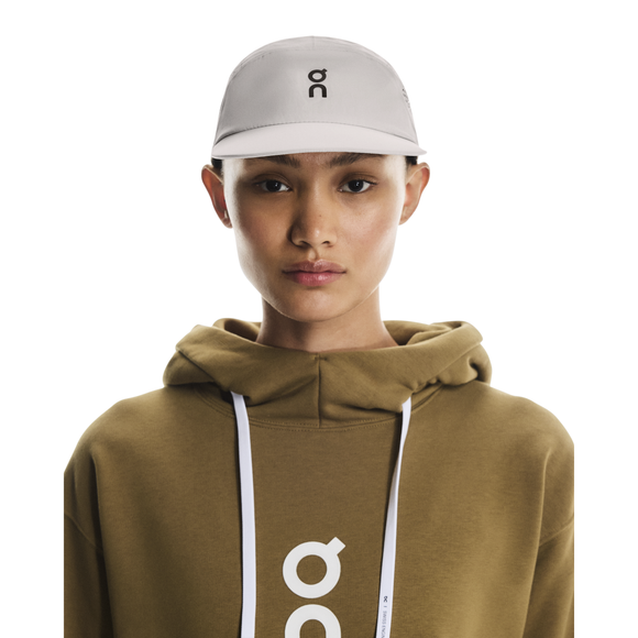 Iført en On Performance Cap i Pearl og en olivengrøn hættetrøje med hvidt logo står personen på en almindelig hvid baggrund og ser direkte ind i kameraet.