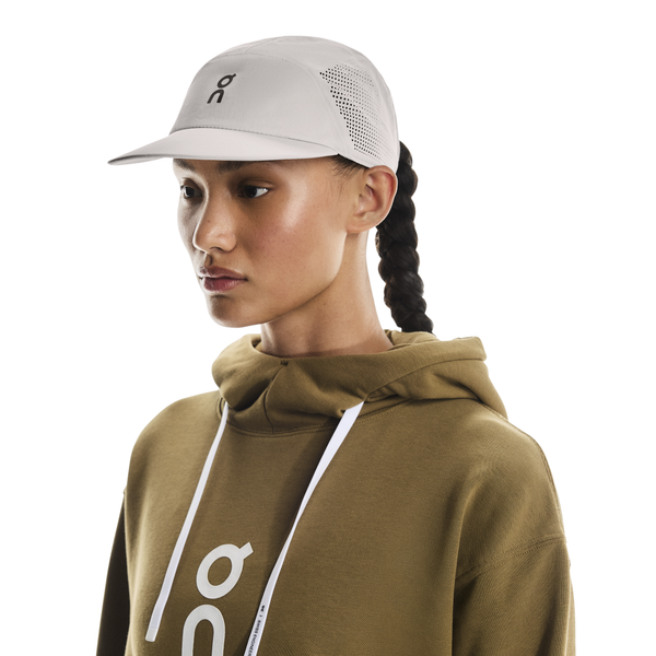 En person i en olivengrøn hættetrøje med hvidt logo og snore bærer On Performance Cap, Unisex, i Pearl, og kigger til siden mod en almindelig hvid baggrund.