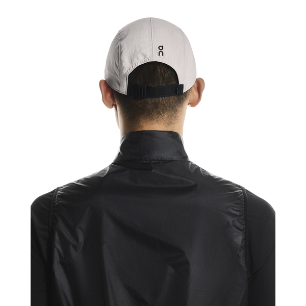 En person ses bagfra iført On Performance Cap, Unisex, Pearl med sort justerbar strop og en sort jakke med høj krave på en almindelig hvid baggrund.