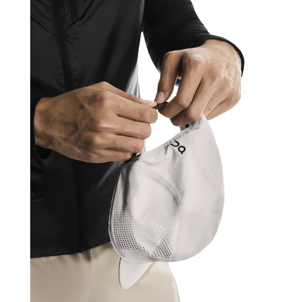 En person i sort jakke og beige bukser folder On Performance Cap, Unisex, Pearl - en let kasket med ventilationshuller - og holder den med begge hænder mod en almindelig hvid baggrund.