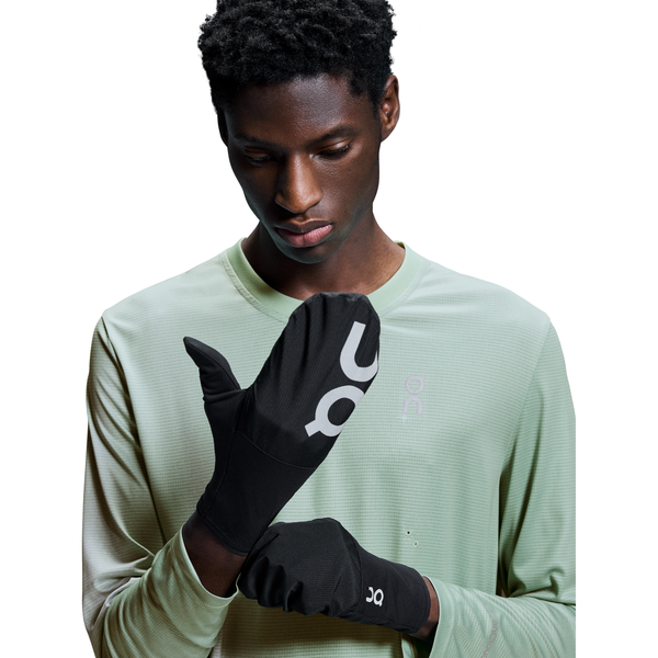 En person i en lysegrøn langærmet skjorte kigger ned, mens han justerer et par sorte Core Glove, Unisex-handsker fra On med et hvidt logo på en hvid baggrund.