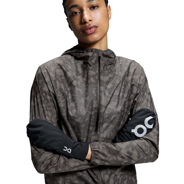 En person iført en mønstret jakke med lynlås og hætte og On's unisex Core Glove i sort med et hvidt OC-logo på hver handske. Jakken har et diskret abstrakt print i jordfarver. Personen står med armene over kors.