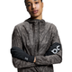 En person iført en mønstret jakke med lynlås og hætte og On's unisex Core Glove i sort med et hvidt OC-logo på hver handske. Jakken har et diskret abstrakt print i jordfarver. Personen står med armene over kors.
