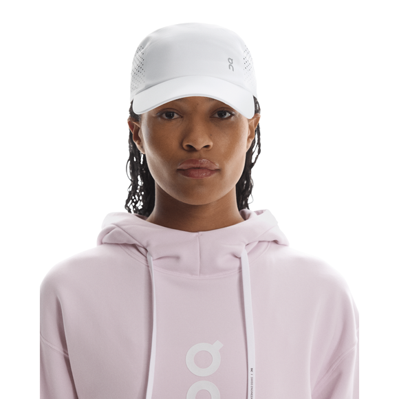 En person iført en On Lightweight Cap, Unisex, i hvid og en lyserød hættetrøje med et hvidt logo står med front mod en almindelig hvid baggrund.