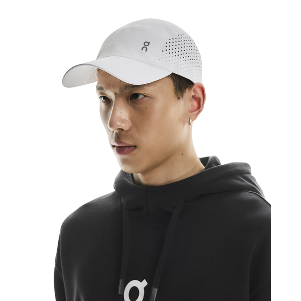 En person iført On Lightweight Cap, Unisex, White med perforeret side og sort hoodie kigger let til højre foran en ensfarvet hvid baggrund. Kasketten er let og ideel til solbeskyttelse på aktive dage.