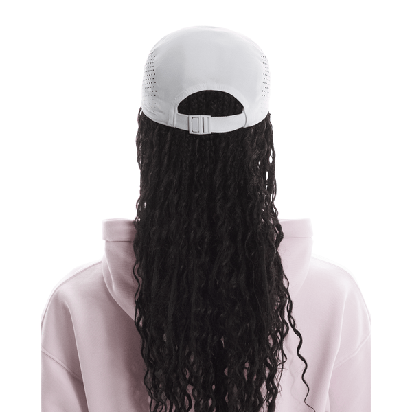 Person med langt, krøllet sort hår bærer en On Lightweight Cap, Unisex, Hvid, bagudvendt og en lyserød hættetrøje. Billedet er taget bagfra mod en hvid baggrund - ideelt til solbeskyttelse på lyse dage.