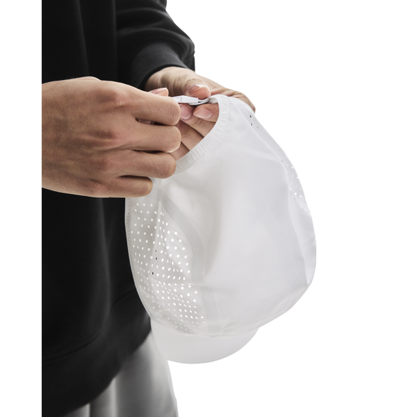 En person i sort langærmet skjorte og lyse bukser justerer bagstroppen på On Lightweight Cap, Unisex, White. Den ensfarvede hvide baggrund fremhæver kaskettens moderne design og solbeskyttelse.