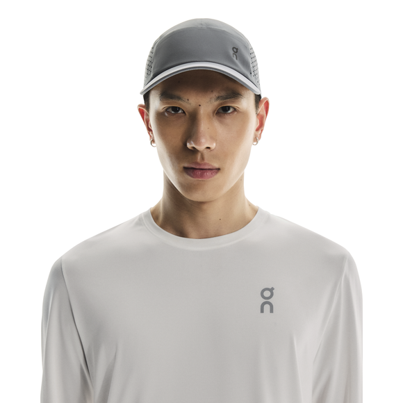 En person iført en On Lightweight Cap, Unisex, Rock i lysegrå og en hvid langærmet skjorte med et lille logo på brystet står på en almindelig hvid baggrund.