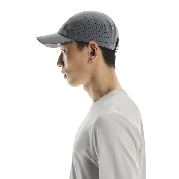 En person med kort mørkt hår, i profil mod en hvid baggrund, bærer en grå "Lightweight Cap, Unisex, Rock" fra On og en lysegrå langærmet skjorte.