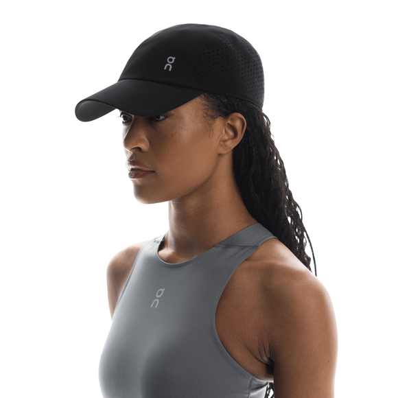 En kvinde iført On Lightweight Cap, Unisex, Black og en grå ærmeløs sportstop står mod en hvid baggrund, vendt mod venstre. Hendes hår er sat op, og hun har et neutralt udtryk.