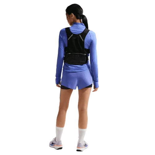 En kvinde med hestehale står med ansigtet vendt mod en hvid baggrund iført Nike Swift UV Half Zip Running Top til kvinder i safir/reflekterende sølv, matchende blå shorts, sort svedtransporterende løbetop, hvide sokker og løbesko.