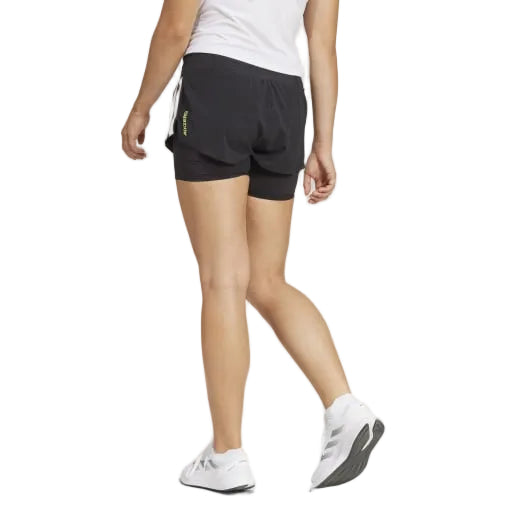 En person i hvid t-shirt, Adidas Adizero 2I1 Shorts Dame Black og hvide sneakers går væk fra kameraet på en almindelig lys baggrund med venstre arm afslappet ved siden af sig, klar til at løbe.