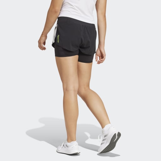 En person i hvid t-shirt, Adidas Adizero 2I1 Shorts Dame Black og hvide sneakers går væk fra kameraet på en almindelig lys baggrund med venstre arm afslappet ved siden af sig, klar til at løbe.