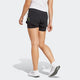 En person i hvid t-shirt, Adidas Adizero 2I1 Shorts Dame Black og hvide sneakers går væk fra kameraet på en almindelig lys baggrund med venstre arm afslappet ved siden af sig, klar til at løbe.