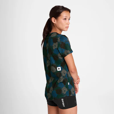En kvinde står sidelæns og kigger sig over skulderen. Hun er iført Saysky W Flower Combat T-Shirt, Dame, Blue Aop med et blåt og grønt blomstermønster og sorte SATISFY-shorts på en almindelig hvid baggrund.