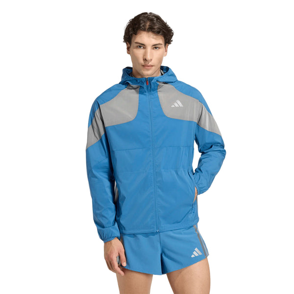 En mand står med front mod en hvid baggrund, iført en Adidas Adizero Hakone Ekiden Jacket i Duinn/Sliver med matchende blå shorts og venstre hånd i shortslommen.