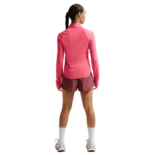 En person med flettet hår står med ryggen til og er iført en Nike Swift UV Half Zip Running Top til kvinder i Sea Coral/Reflective Silver, rødbrune sportsshorts, hvide sokker og hvide sneakers på en almindelig lys baggrund.