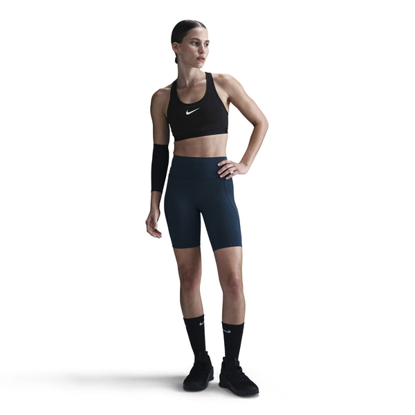En kvinde står på en hvid baggrund iført en sort sports-bh og Nike Dame Medium-Support High-Waisted 8" Biker Shorts i Black/Anthracite/Dk Smoke Grey, parret med sorte knæstrømper, sorte sneakers og et sort ærme på venstre arm.