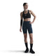En kvinde står på en hvid baggrund iført en sort sports-bh og Nike Dame Medium-Support High-Waisted 8" Biker Shorts i Black/Anthracite/Dk Smoke Grey, parret med sorte knæstrømper, sorte sneakers og et sort ærme på venstre arm.