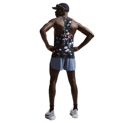 En mand står med hænderne på hofterne, iført Nike Aeroswift Singlet Aop, Herre, Black/Laser Orange - perfekt svedtransporterende træningstøj - sammen med blå shorts, hvide løbesko og en sort kasket, og kigger væk fra kameraet.