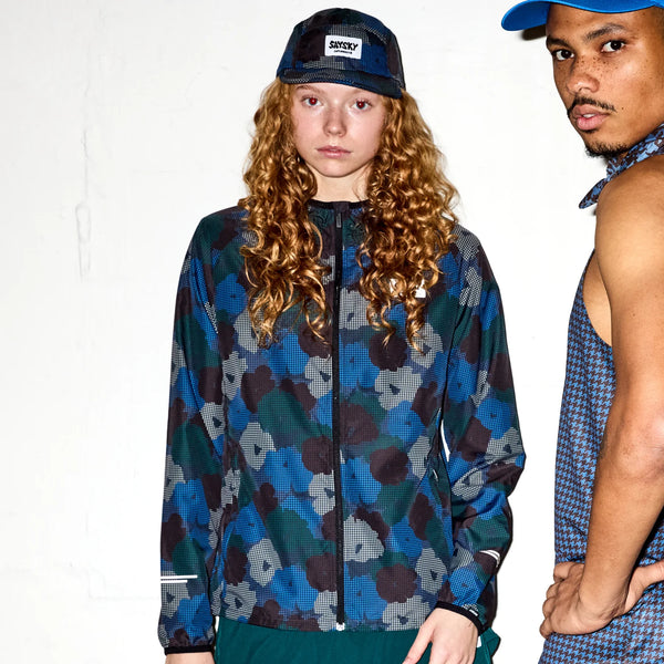 To personer poserer mod en hvid baggrund. Personen til venstre har en mønstret jakke på og en Saysky Flower Combat Cap 103, Unisex, Blue Aop. Personen til højre bærer en ærmeløs skjorte med blomsterprint og en hat; begge vender mod kameraet.