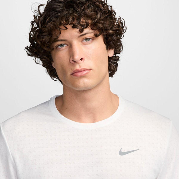 En ung mand med krøllet brunt hår og blå øjne iført Nike Miler Short-Sleeve Running Top, Herre, Summit White/Reflective Silver, ser direkte ind i kameraet med et neutralt udtryk mod en almindelig lys baggrund.