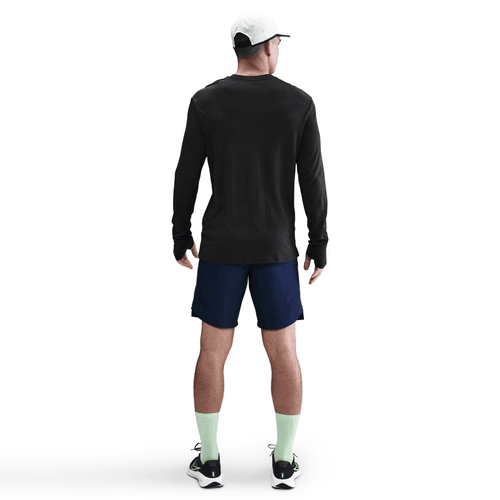 En person står med ansigtet vendt mod en hvid baggrund iført en Nike Dri-FIT ADV Wool langærmet løbetop til mænd i sort/antrazit, hvid kasket, mørkeblå shorts, lysegrønne sokker og sorte sportssko.