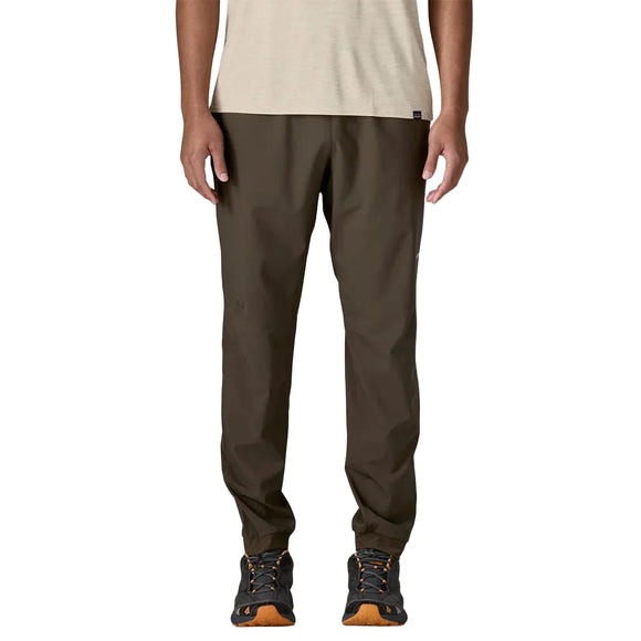 En mand står foran en ensfarvet baggrund iført Patagonia Terrebonne Joggers i Otter Brown. Han kombinerer dem med en lys beige kortærmet skjorte og sorte vandresko med orange detaljer. Billedet er beskåret lige under skuldrene.