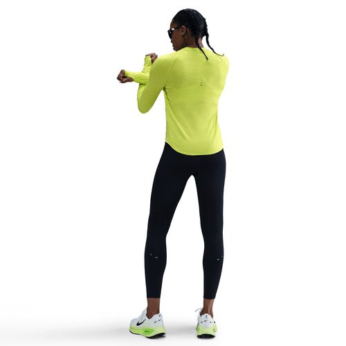 En kvinde i en knaldgul Nike Dri-FIT UV Long-Sleeve Crew-Neck Running Top (Cyber/Htr/Reflective Silv), sorte leggings, hvide sneakers og solbriller står og strækker sin venstre arm ud over kroppen mod en almindelig hvid baggrund.
