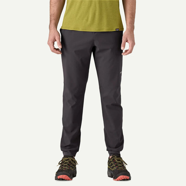 En person iført en lysegrøn kortærmet skjorte, Patagonia Terrebonne Joggers, Herre, Black, og sorte trail-løbesko med orange og gule detaljer står på en almindelig hvid baggrund.