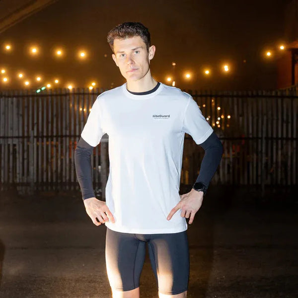 En mand står udendørs om aftenen iført LiiteGuard Ground-Tech T-shirt, Herre, White over en sort langærmet top og sorte shorts, med lysende lyskæder i baggrunden og hænderne på hofterne.
